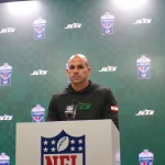 FootballR - NFL - Robert Saleh steht an einem Podium mit einem NFL-Logo und trägt einen schwarzen Kapuzenpullover mit einem grünen „Jets“-Logo. Der Hintergrund ist eine grüne Wand mit den Logos „Jets“ und „NFL London“, was an eine automatisch arrangierte Pressekonferenz oder ein Sportereignis erinnert. Diese Beschreibung wurde automatisch generiert.
