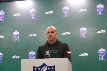 FootballR - NFL - Robert Saleh steht an einem Podium mit einem NFL-Logo und trägt einen schwarzen Kapuzenpullover mit einem grünen „Jets“-Logo. Der Hintergrund ist eine grüne Wand mit den Logos „Jets“ und „NFL London“, was an eine automatisch arrangierte Pressekonferenz oder ein Sportereignis erinnert. Diese Beschreibung wurde automatisch generiert.