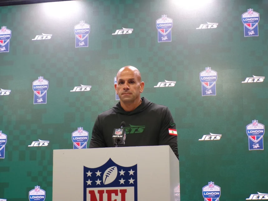 FootballR - NFL - Robert Saleh steht an einem Podium mit einem NFL-Logo und trägt einen schwarzen Kapuzenpullover mit einem grünen „Jets“-Logo. Der Hintergrund ist eine grüne Wand mit den Logos „Jets“ und „NFL London“, was an eine automatisch arrangierte Pressekonferenz oder ein Sportereignis erinnert. Diese Beschreibung wurde automatisch generiert.