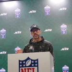 FootballR - NFL - Aaron Rodgers - Eine Person steht an einem Podium mit einem NFL-Logo und trägt einen Camouflage-Kapuzenpulli und eine schwarze Kappe. Dahinter ist ein grün karierter Hintergrund mit Logos der New York Jets und der NFL London zu sehen. Sie scheint einen Entwurf zu präsentieren, mit aufmerksamem Auftreten. Diese Beschreibung wurde automatisch generiert.