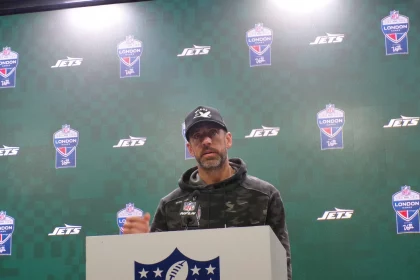 FootballR - NFL - Aaron Rodgers - Eine Person steht an einem Podium mit einem NFL-Logo und trägt einen Camouflage-Kapuzenpulli und eine schwarze Kappe. Dahinter ist ein grün karierter Hintergrund mit Logos der New York Jets und der NFL London zu sehen. Sie scheint einen Entwurf zu präsentieren, mit aufmerksamem Auftreten. Diese Beschreibung wurde automatisch generiert.