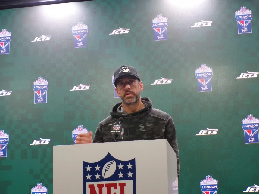 FootballR - NFL - Aaron Rodgers - Eine Person steht an einem Podium mit einem NFL-Logo und trägt einen Camouflage-Kapuzenpulli und eine schwarze Kappe. Dahinter ist ein grün karierter Hintergrund mit Logos der New York Jets und der NFL London zu sehen. Sie scheint einen Entwurf zu präsentieren, mit aufmerksamem Auftreten. Diese Beschreibung wurde automatisch generiert.