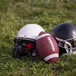 FootballR - NFL - Zwei American-Football-Helme, einer weiß und einer schwarz, liegen neben einem Football auf einem Rasenfeld. Der Ball steht aufrecht zwischen den Helmen auf dem hellgrünen Gras und lädt Sportfans und NFL-Wetten-Enthusiasten ein, diesen sonnigen Tag voller Nervenkitzel im Freien zu genießen. Diese Beschreibung wurde automatisch generiert.