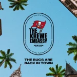Von unten rahmen Gebäude im europäischen Stil und Palmen einen klaren blauen Himmel ein. In der Mitte befindet sich „The Krewe Embassy“, geschmückt mit einem Piraten-Emblem, das es als offizielles Heimstadion der Tampa Bay Buccaneers in München kennzeichnet. Darunter steht in fettem Text: „Die Bucs sind zurück in der Stadt.“ Diese Beschreibung wurde mit der FootballR KI automatisch generiert.