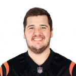 Ein Mann mit kurzen dunklen Haaren und Bart, möglicherweise Ben Brown, lächelt in die Kamera. Er trägt ein schwarzes Sporttrikot mit orangefarbenen Streifen auf den Schultern und einem NFL-Logo am Kragen. Der Hintergrund ist komplett schwarz. Diese Beschreibung wurde mit der FootballR KI automatisch generiert.