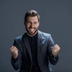 FootballR - NFL - Ein Mann in grauem Anzug und dunklem gepunktetem Hemd steht vor einem einfarbig grauen Hintergrund, lächelt breit und ballt aufgeregt die Fäuste, als hätte er gerade das perfekte DAZN Abo ergattert. Sein Haar ist ordentlich gestylt, er hat Stoppeln im Gesicht und ein buntes Einstecktuch peppt sein Jackett auf. Diese Beschreibung wurde automatisch generiert.