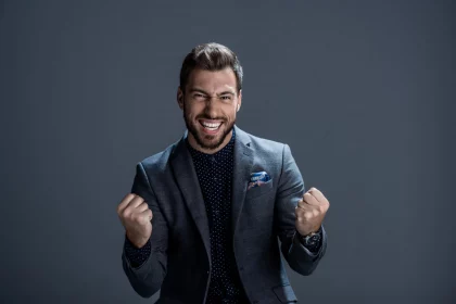 FootballR - NFL - Ein Mann in grauem Anzug und dunklem gepunktetem Hemd steht vor einem einfarbig grauen Hintergrund, lächelt breit und ballt aufgeregt die Fäuste, als hätte er gerade das perfekte DAZN Abo ergattert. Sein Haar ist ordentlich gestylt, er hat Stoppeln im Gesicht und ein buntes Einstecktuch peppt sein Jackett auf. Diese Beschreibung wurde automatisch generiert.