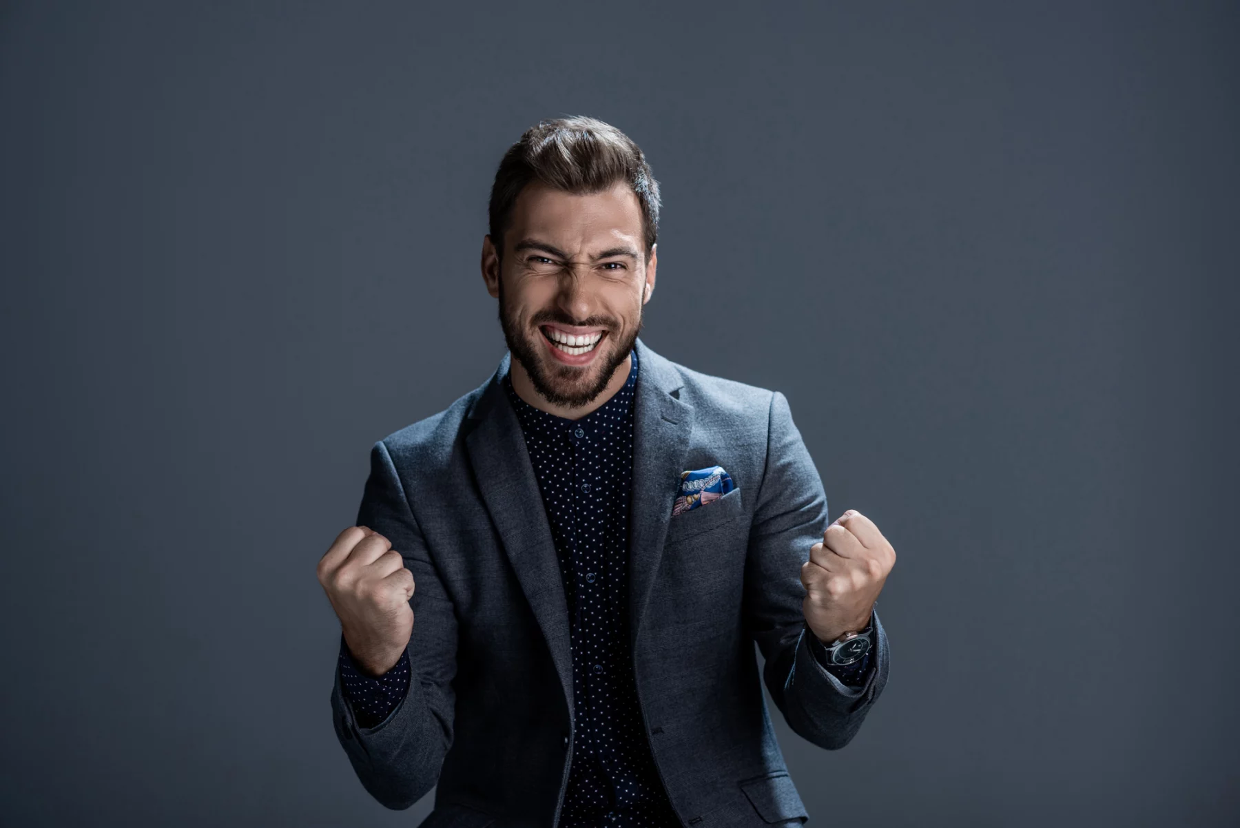 FootballR - NFL - Ein Mann in grauem Anzug und dunklem gepunktetem Hemd steht vor einem einfarbig grauen Hintergrund, lächelt breit und ballt aufgeregt die Fäuste, als hätte er gerade das perfekte DAZN Abo ergattert. Sein Haar ist ordentlich gestylt, er hat Stoppeln im Gesicht und ein buntes Einstecktuch peppt sein Jackett auf. Diese Beschreibung wurde automatisch generiert.
