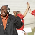 FootballR - NFL - Ein älterer Mann in schwarzer Jacke und mit Kopfhörern, vermutlich ein Trainer, jubelt leidenschaftlich mit erhobener Faust. Im Hintergrund jubeln zwei Footballspieler in roten Trikots, einer davon mit der Nummer 27, mit erhobenen Armen und gefalteten Händen in einem Gebäude, das wie ein Freiluftstadion für American Football aussieht. Diese Beschreibung wurde automatisch generiert.