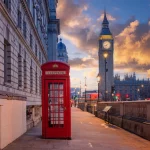 FootballR - NFL - London Flag Football - Eine rote britische Telefonzelle steht bei Sonnenuntergang in einer ruhigen Straße. Ihr ikonisches Design (Entwurf) bildet einen schönen Kontrast zu Big Ben und den Houses of Parliament. Dramatische Wolken füllen den Himmel, während Straßenlaternen im Vintage-Stil historische Gebäude beleuchten und diesen Moment konservieren, als ob er automatisch gespeichert wäre. Diese Beschreibung wurde automatisch generiert.