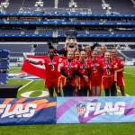 FootballR - AFL - Eine Gruppe von Frauen in roten Sportuniformen steht lächelnd auf einem Fußballfeld in London und hält eine dänische Flagge in den Händen. Hinter ihnen steht ein Maskottchen in schwarzer Uniform und mit Hut. NFL-FLAG-Banner sind deutlich zu sehen, im Hintergrund leuchtet ein leeres blaues Stadion. Diese Beschreibung wurde automatisch generiert.
