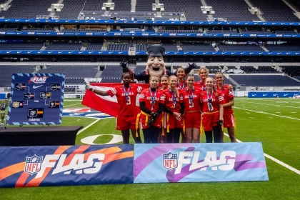 FootballR - AFL - Eine Gruppe von Frauen in roten Sportuniformen steht lächelnd auf einem Fußballfeld in London und hält eine dänische Flagge in den Händen. Hinter ihnen steht ein Maskottchen in schwarzer Uniform und mit Hut. NFL-FLAG-Banner sind deutlich zu sehen, im Hintergrund leuchtet ein leeres blaues Stadion. Diese Beschreibung wurde automatisch generiert.