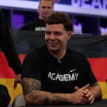 FootballR - NFL - Kevin Bentin, mit kurzen, lockigen dunklen Haaren, sitzt lächelnd an einem Tisch und trägt ein schwarzes Nike-Shirt mit der Aufschrift „ACADEMY“. Im Hintergrund wird stolz eine deutsche Flagge hochgehalten. Rechts auf dem Tisch liegt ein schwarz-weißer Helm. Andere Menschen und verschwommene Schilder füllen die Szene hinter ihm. Diese Beschreibung wurde automatisch generiert.