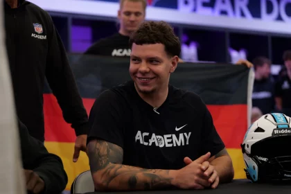 FootballR - NFL - Kevin Bentin, mit kurzen, lockigen dunklen Haaren, sitzt lächelnd an einem Tisch und trägt ein schwarzes Nike-Shirt mit der Aufschrift „ACADEMY“. Im Hintergrund wird stolz eine deutsche Flagge hochgehalten. Rechts auf dem Tisch liegt ein schwarz-weißer Helm. Andere Menschen und verschwommene Schilder füllen die Szene hinter ihm. Diese Beschreibung wurde automatisch generiert.