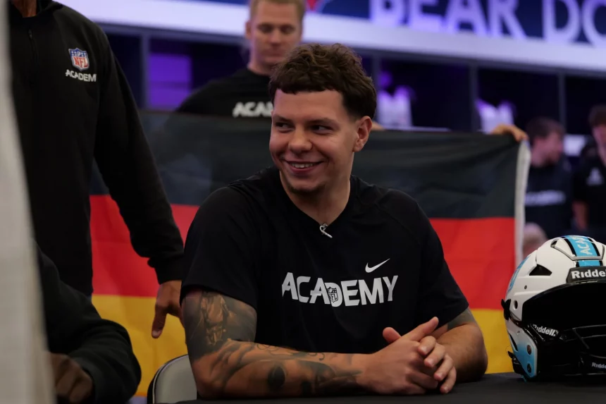 FootballR - NFL - Kevin Bentin, mit kurzen, lockigen dunklen Haaren, sitzt lächelnd an einem Tisch und trägt ein schwarzes Nike-Shirt mit der Aufschrift „ACADEMY“. Im Hintergrund wird stolz eine deutsche Flagge hochgehalten. Rechts auf dem Tisch liegt ein schwarz-weißer Helm. Andere Menschen und verschwommene Schilder füllen die Szene hinter ihm. Diese Beschreibung wurde automatisch generiert.