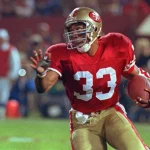 FootballR - NFL Hall of Fame - Footballspieler Anderson, mit der roten Trikotnummer 33 und einem goldenen Helm, stürmt über das Feld. Roger Craig trägt goldene Hosen und schwarze Stollenschuhe und hat seine rechte Hand ausgestreckt. Zuschauer und Teammitglieder verschwimmen unter den Stadionlichtern, während er entschlossen voranschreitet. Diese Beschreibung wurde automatisch generiert.