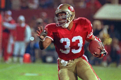 FootballR - NFL Hall of Fame - Footballspieler Anderson, mit der roten Trikotnummer 33 und einem goldenen Helm, stürmt über das Feld. Roger Craig trägt goldene Hosen und schwarze Stollenschuhe und hat seine rechte Hand ausgestreckt. Zuschauer und Teammitglieder verschwimmen unter den Stadionlichtern, während er entschlossen voranschreitet. Diese Beschreibung wurde automatisch generiert.