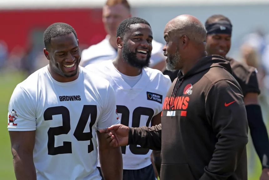 FootballR - NFL - Drei Männer in Trainingskleidung der Cleveland Browns stehen auf einem Footballfeld. Zwei Spieler, darunter Nick Chubb mit der Trikotnummer 24, lächeln und sprechen mit einem Trainer in einem braunen Kapuzenpullover. Der sonnige Tag und der unscharfe Hintergrund lassen auf eine Übung oder Trainingseinheit schließen. Diese Beschreibung wurde automatisch generiert.