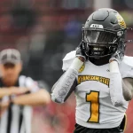 FootballR - NFL - Ein Footballspieler mit schwarzem Helm und weißem Trikot mit der Aufschrift Kennesaw State in Gelb steht nach einem Sensationssieg auf dem Spielfeld, die Hände auf dem Helm. Sein rechter Arm, umwickelt mit weißem Klebeband mit Schriftzug, ist zu sehen. Im Hintergrund signalisiert ein Schiedsrichter mit verschwommenen roten Stadionsitzen, die Libertys Spielfeld umgeben. Diese Beschreibung wurde automatisch generiert.