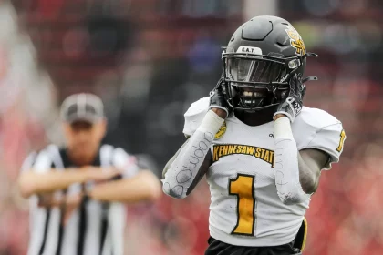 FootballR - NFL - Ein Footballspieler mit schwarzem Helm und weißem Trikot mit der Aufschrift Kennesaw State in Gelb steht nach einem Sensationssieg auf dem Spielfeld, die Hände auf dem Helm. Sein rechter Arm, umwickelt mit weißem Klebeband mit Schriftzug, ist zu sehen. Im Hintergrund signalisiert ein Schiedsrichter mit verschwommenen roten Stadionsitzen, die Libertys Spielfeld umgeben. Diese Beschreibung wurde automatisch generiert.