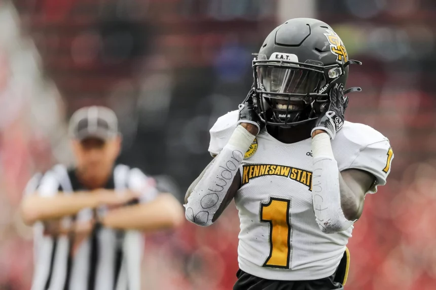 FootballR - NFL - Ein Footballspieler mit schwarzem Helm und weißem Trikot mit der Aufschrift Kennesaw State in Gelb steht nach einem Sensationssieg auf dem Spielfeld, die Hände auf dem Helm. Sein rechter Arm, umwickelt mit weißem Klebeband mit Schriftzug, ist zu sehen. Im Hintergrund signalisiert ein Schiedsrichter mit verschwommenen roten Stadionsitzen, die Libertys Spielfeld umgeben. Diese Beschreibung wurde automatisch generiert.