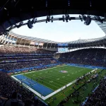 FootballR - NFL - Eine Weitwinkelansicht eines voll besetzten Footballstadions während der RTL NFL 2024. Zu sehen ist ein grünes Feld mit sichtbaren Yard-Linien und Torpfosten. Während eines Spiels sind die Spieler auf dem Feld, während die Zuschauer die Tribünen füllen und große Bildschirme Spielinformationen anzeigen. Das Dach des Stadions ist bei klarem Himmel teilweise sichtbar. Diese Beschreibung wurde automatisch generiert.