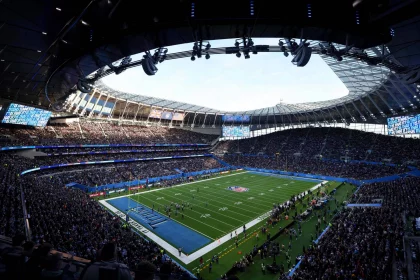 FootballR - NFL - Eine Weitwinkelansicht eines voll besetzten Footballstadions während der RTL NFL 2024. Zu sehen ist ein grünes Feld mit sichtbaren Yard-Linien und Torpfosten. Während eines Spiels sind die Spieler auf dem Feld, während die Zuschauer die Tribünen füllen und große Bildschirme Spielinformationen anzeigen. Das Dach des Stadions ist bei klarem Himmel teilweise sichtbar. Diese Beschreibung wurde automatisch generiert.
