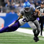 FootballR - NFL Free TV - Amon-Ra St. Brown in grauer Uniform, mit der Nummer 14 und blauem Helm, klammert sich beim Laufen über das Feld an den Ball. Ein Spieler in lila-weißer Uniform versucht, ihn unter den wachsamen Augen eines Schiedsrichters zu tackeln. Verfolgen Sie diese ganze Aufregung live im Free-TV. Diese Beschreibung wurde automatisch generiert.