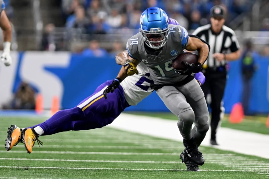 FootballR - NFL Free TV - Amon-Ra St. Brown in grauer Uniform, mit der Nummer 14 und blauem Helm, klammert sich beim Laufen über das Feld an den Ball. Ein Spieler in lila-weißer Uniform versucht, ihn unter den wachsamen Augen eines Schiedsrichters zu tackeln. Verfolgen Sie diese ganze Aufregung live im Free-TV. Diese Beschreibung wurde automatisch generiert.