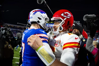FootballR - NFL Awards - Zwei American-Football-Spieler, einer in der blauen Uniform der Buffalo Bills und der andere in der roten Uniform der Kansas City Chiefs, umarmen sich während der NFL Awards auf dem Spielfeld. Sie sind von Kameras und Mikrofonen umgeben. Im Hintergrund ist das Stadion zu sehen, und andere beobachten das Zusammenspiel. Diese Beschreibung wurde automatisch generiert.