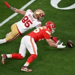 FootballR - NFL - In Woche 7 treffen die Footballspieler der Chiefs und 49ers erneut auf einem grünen Feld aufeinander. Der Spieler in der rot-weißen Uniform greift nach dem Ball, während die Nummer 85 in Rot und Gold nach vorne hechtet. Beide sind intensiv auf den Ball konzentriert und fangen damit die Essenz des Wettkampfs ein. Diese Beschreibung wurde automatisch generiert.