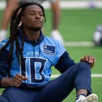 FootballR - NFL - DeAndre Hopkins mit langen Dreadlocks sitzt auf dem Feld und macht Dehnübungen. Er trägt ein hellblaues Trikot mit der Nummer 10 und dunkle Hosen und wirkt konzentriert und entspannt, während er sich auf das Training vorbereitet. In der Nähe liegt ein Helm mit dem Logo der Kansas City Chiefs neben einer Dose auf dem Rasen. Diese Beschreibung wurde automatisch generiert.