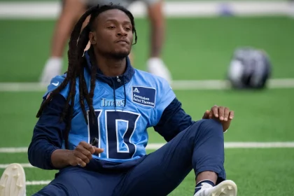 FootballR - NFL - DeAndre Hopkins mit langen Dreadlocks sitzt auf dem Feld und macht Dehnübungen. Er trägt ein hellblaues Trikot mit der Nummer 10 und dunkle Hosen und wirkt konzentriert und entspannt, während er sich auf das Training vorbereitet. In der Nähe liegt ein Helm mit dem Logo der Kansas City Chiefs neben einer Dose auf dem Rasen. Diese Beschreibung wurde automatisch generiert.