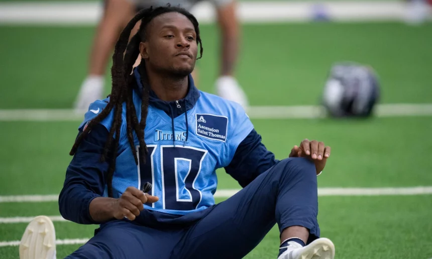 FootballR - NFL - DeAndre Hopkins mit langen Dreadlocks sitzt auf dem Feld und macht Dehnübungen. Er trägt ein hellblaues Trikot mit der Nummer 10 und dunkle Hosen und wirkt konzentriert und entspannt, während er sich auf das Training vorbereitet. In der Nähe liegt ein Helm mit dem Logo der Kansas City Chiefs neben einer Dose auf dem Rasen. Diese Beschreibung wurde automatisch generiert.