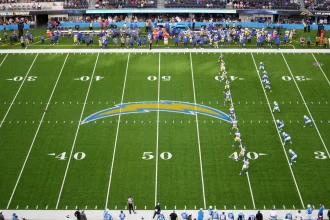 FootballR Onside Kick im Rahmen des dynamischen Kickoff - NFL Kickoff Regel - In einem NFL-Stadion entfaltet sich ein spannendes Footballspiel, während sich zwei Teams auf dem Spielfeld aufstellen. Das Team in blau-weißen Uniformen bereitet sich auf die Kickoff-Regel vor und versucht, sich einen frühen Vorteil zu verschaffen. Yard-Linien und ein auffälliges Teamlogo markieren das Spielfeld, während Zuschauer von den Tribünen jubeln und Mitarbeiter an den Seitenlinien beschäftigt sind. Diese Beschreibung wurde automatisch generiert.