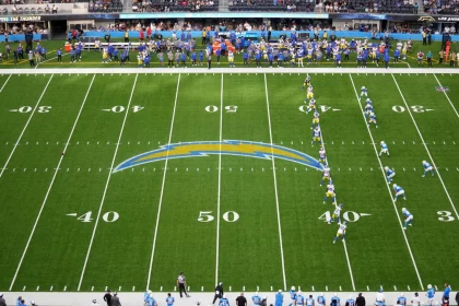 FootballR Onside Kick im Rahmen des dynamischen Kickoff - NFL Kickoff Regel - In einem NFL-Stadion entfaltet sich ein spannendes Footballspiel, während sich zwei Teams auf dem Spielfeld aufstellen. Das Team in blau-weißen Uniformen bereitet sich auf die Kickoff-Regel vor und versucht, sich einen frühen Vorteil zu verschaffen. Yard-Linien und ein auffälliges Teamlogo markieren das Spielfeld, während Zuschauer von den Tribünen jubeln und Mitarbeiter an den Seitenlinien beschäftigt sind. Diese Beschreibung wurde automatisch generiert.