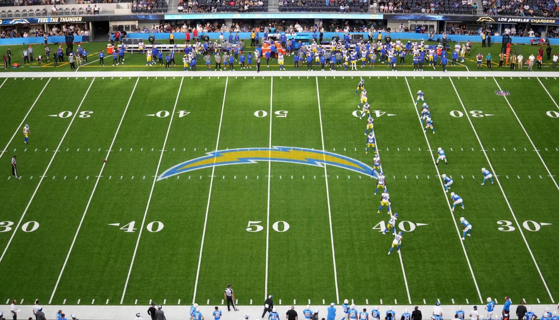 FootballR Onside Kick im Rahmen des dynamischen Kickoff - NFL Kickoff Regel - In einem NFL-Stadion entfaltet sich ein spannendes Footballspiel, während sich zwei Teams auf dem Spielfeld aufstellen. Das Team in blau-weißen Uniformen bereitet sich auf die Kickoff-Regel vor und versucht, sich einen frühen Vorteil zu verschaffen. Yard-Linien und ein auffälliges Teamlogo markieren das Spielfeld, während Zuschauer von den Tribünen jubeln und Mitarbeiter an den Seitenlinien beschäftigt sind. Diese Beschreibung wurde automatisch generiert.