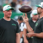 FootballR - NFL - Zwei Personen stehen auf dem Footballfeld, gekleidet in New York Jets-Ausrüstung. Aaron Rodgers rechts wirft einen Football in die Luft, vielleicht inspiriert von Nathaniel Hackett. Beide tragen Jets-Kappen und schwarze Shirts, im Hintergrund sind eine verschwommene Menschenmenge und grüne Stadionsitze zu sehen. Diese Beschreibung wurde automatisch generiert.