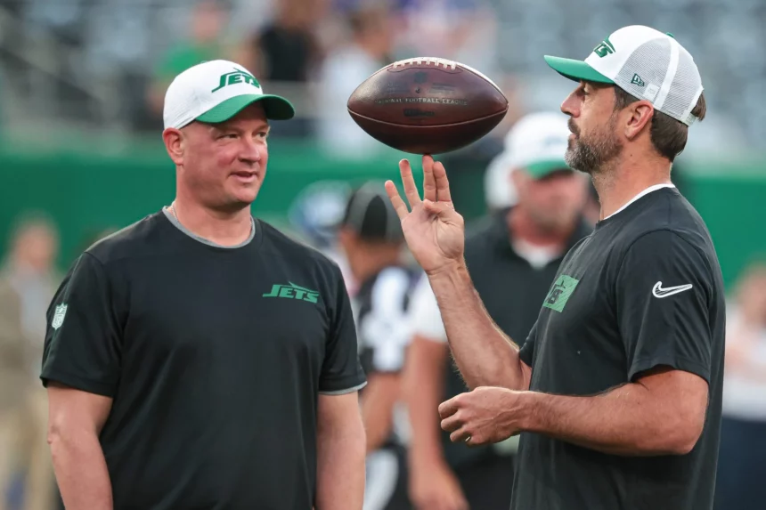 FootballR - NFL - Zwei Personen stehen auf dem Footballfeld, gekleidet in New York Jets-Ausrüstung. Aaron Rodgers rechts wirft einen Football in die Luft, vielleicht inspiriert von Nathaniel Hackett. Beide tragen Jets-Kappen und schwarze Shirts, im Hintergrund sind eine verschwommene Menschenmenge und grüne Stadionsitze zu sehen. Diese Beschreibung wurde automatisch generiert.