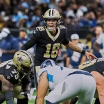 FootballR - NFL - Ein Quarterback in schwarz-goldener Uniform, Nummer 18, ruft auf dem Feld einen Spielzug aus, was an Spencer Rattler erinnert. Zwei Teamkollegen in ähnlichen Uniformen hocken vor einem Spieler in weiß-blauer Uniform. Im Hintergrund sind Zuschauer verschwommen zu sehen. Diese Beschreibung wurde automatisch generiert.