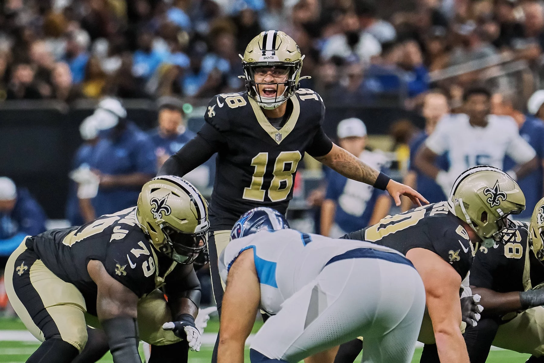 FootballR - NFL - Ein Quarterback in schwarz-goldener Uniform, Nummer 18, ruft auf dem Feld einen Spielzug aus, was an Spencer Rattler erinnert. Zwei Teamkollegen in ähnlichen Uniformen hocken vor einem Spieler in weiß-blauer Uniform. Im Hintergrund sind Zuschauer verschwommen zu sehen. Diese Beschreibung wurde automatisch generiert.