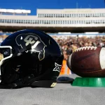 Nikhil Webb Walker - Ein schwarzer Footballhelm mit einem goldenen Colorado Buffaloes Logo steht auf einem Tisch neben einem Football und repräsentiert stolz Colorado. Der Hintergrund zeigt eine große, verschwommene Menschenmenge in einem Stadion an einem sonnigen Tag mit klarem blauen Himmel darüber. Diese Beschreibung wurde mit der FootballR KI automatisch generiert.