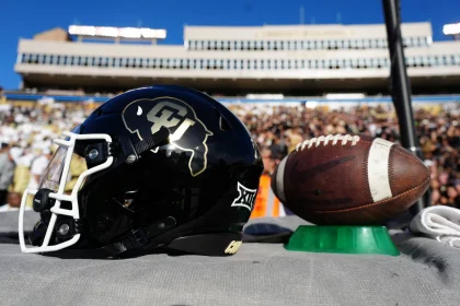 Nikhil Webb Walker - Ein schwarzer Footballhelm mit einem goldenen Colorado Buffaloes Logo steht auf einem Tisch neben einem Football und repräsentiert stolz Colorado. Der Hintergrund zeigt eine große, verschwommene Menschenmenge in einem Stadion an einem sonnigen Tag mit klarem blauen Himmel darüber. Diese Beschreibung wurde mit der FootballR KI automatisch generiert.