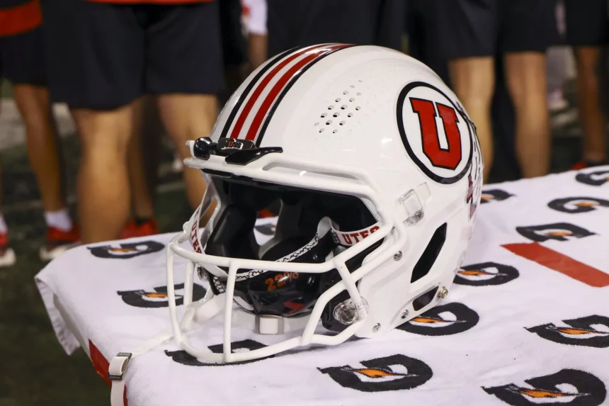 FootballR - College Football - Ein weißer Footballhelm mit rotem „U“-Logo und roten Streifen liegt auf einem Handtuch mit Gatorade-Aufschrift. Er steht auf einem Tisch, in dessen Nähe sich Menschen in Sportshorts aufhalten, die vielleicht darauf warten, dass Utahs Quarterback Cam Rising die nächste Saison mit Elan anführt. Diese Beschreibung wurde automatisch generiert.