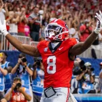 FootballR - NFL - Ein Footballspieler in einem roten Trikot mit der Nummer 8, das an den Wide Receiver der Georgia Bulldogs, Colbie Young, erinnert, jubelt mit erhobenen Armen in einem Stadion. Die jubelnde Menge im Hintergrund winkt wild mit den Händen, und Fotografen am Spielfeldrand halten den denkwürdigen Moment eifrig fest. Diese Beschreibung wurde automatisch generiert.