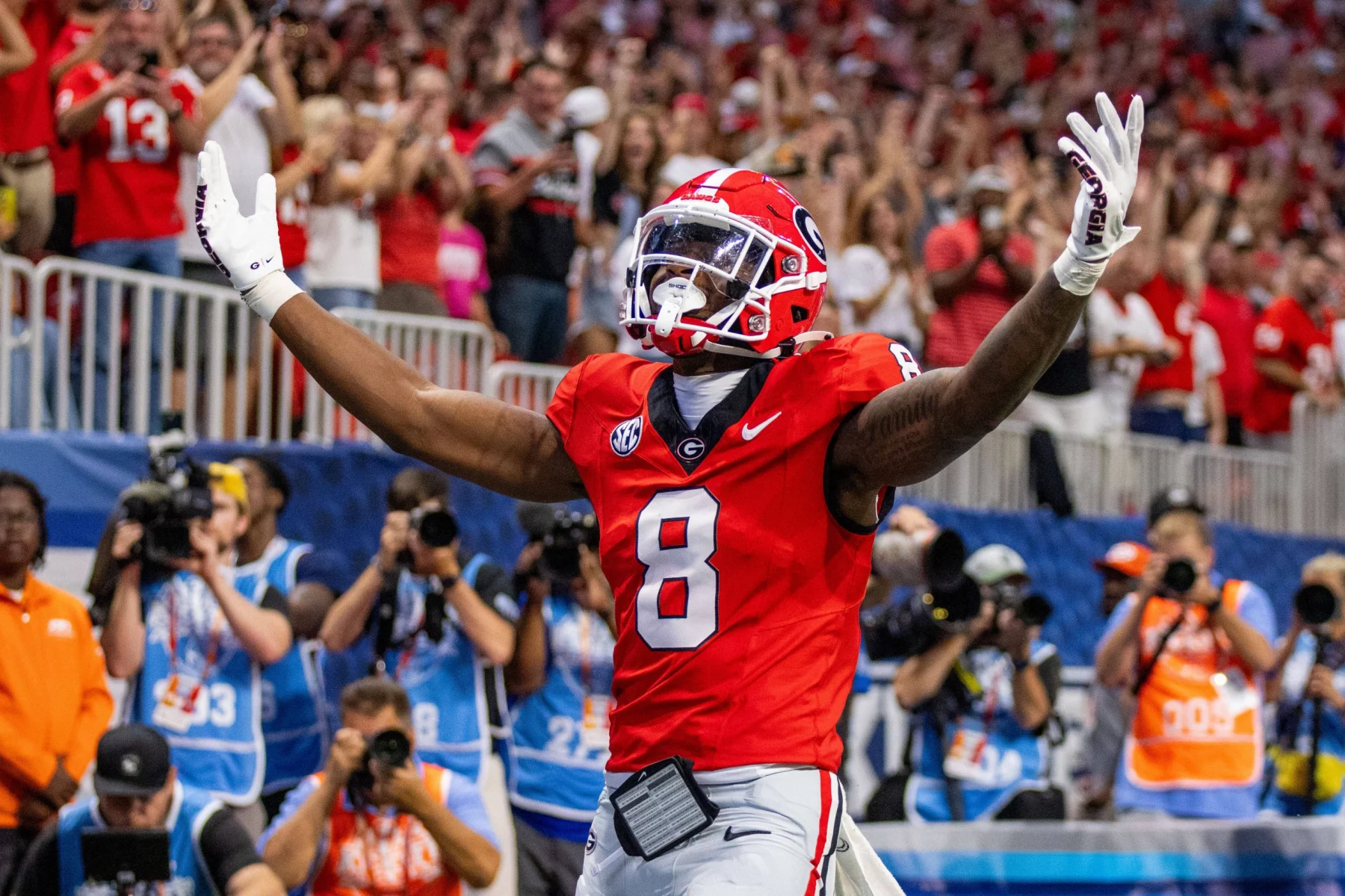 FootballR - NFL - Ein Footballspieler in einem roten Trikot mit der Nummer 8, das an den Wide Receiver der Georgia Bulldogs, Colbie Young, erinnert, jubelt mit erhobenen Armen in einem Stadion. Die jubelnde Menge im Hintergrund winkt wild mit den Händen, und Fotografen am Spielfeldrand halten den denkwürdigen Moment eifrig fest. Diese Beschreibung wurde automatisch generiert.