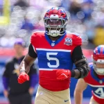 FootballR - NFL - Ein Footballspieler, der an Kayvon Thibodeaux erinnert, trägt eine rot-weiß-blaue Uniform mit der Nummer 5 und sprintet über das Feld. Er trägt einen Helm mit Visier und eine Armschiene, während im Hintergrund ein weiterer Spieler mit der Nummer 41 zu sehen ist. Die Szene im Freien zeigt eine verschwommene Menschenmenge und Feldausrüstung. Diese Beschreibung wurde automatisch generiert.