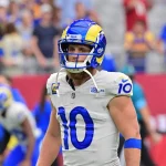 FootballR - NFL - Ein Footballspieler, Cooper Kupp, steht in der Uniform der Los Angeles Rams auf dem Feld. Er trägt einen blau-gelben Helm und ein weißes Trikot mit der Nummer 10. Verschwommene Zuschauer und Teamkollegen in ähnlicher Kleidung füllen den Hintergrund und fangen die Intensität des Spiels ein. Diese Beschreibung wurde automatisch generiert.