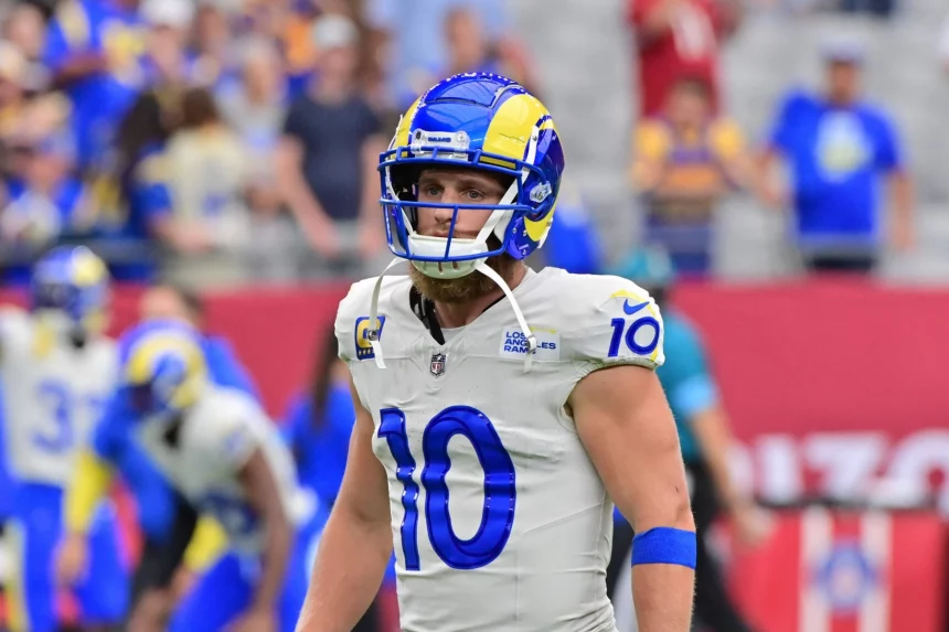 FootballR - NFL - Ein Footballspieler, Cooper Kupp, steht in der Uniform der Los Angeles Rams auf dem Feld. Er trägt einen blau-gelben Helm und ein weißes Trikot mit der Nummer 10. Verschwommene Zuschauer und Teamkollegen in ähnlicher Kleidung füllen den Hintergrund und fangen die Intensität des Spiels ein. Diese Beschreibung wurde automatisch generiert.