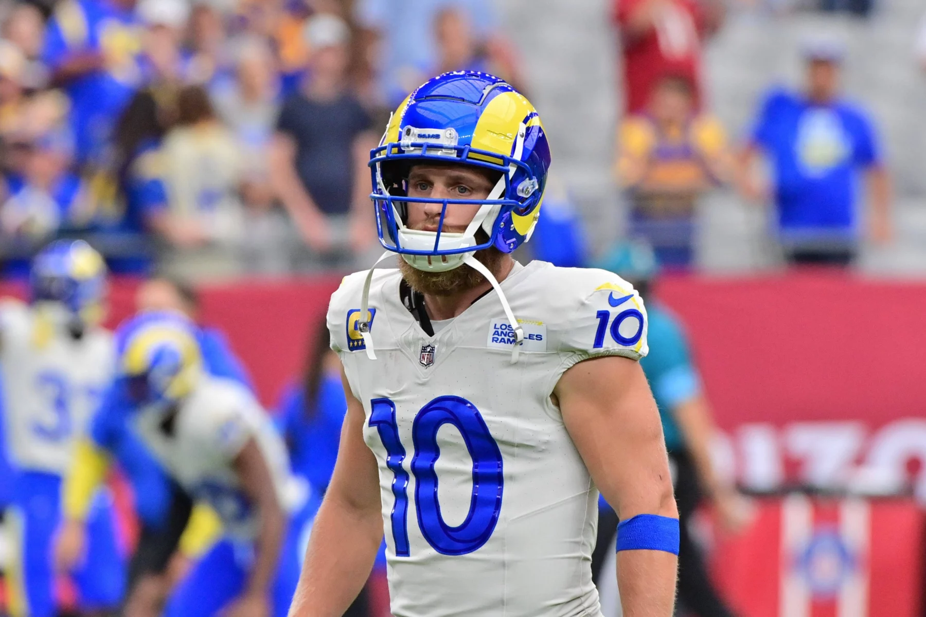 FootballR - NFL - Ein Footballspieler, Cooper Kupp, steht in der Uniform der Los Angeles Rams auf dem Feld. Er trägt einen blau-gelben Helm und ein weißes Trikot mit der Nummer 10. Verschwommene Zuschauer und Teamkollegen in ähnlicher Kleidung füllen den Hintergrund und fangen die Intensität des Spiels ein. Diese Beschreibung wurde automatisch generiert.